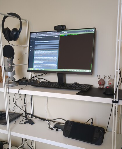 image of a steam deck, USB-C dock, external monitor, and cable spaghetti - all squished onto a cluttered and unstable bookshelf, in lieu of a desk. 